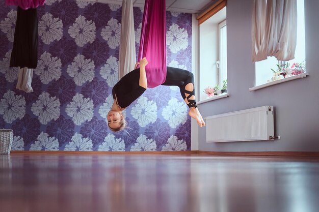 Mujer joven haciendo práctica de yoga aérea en hamaca morada en el gimnasio.