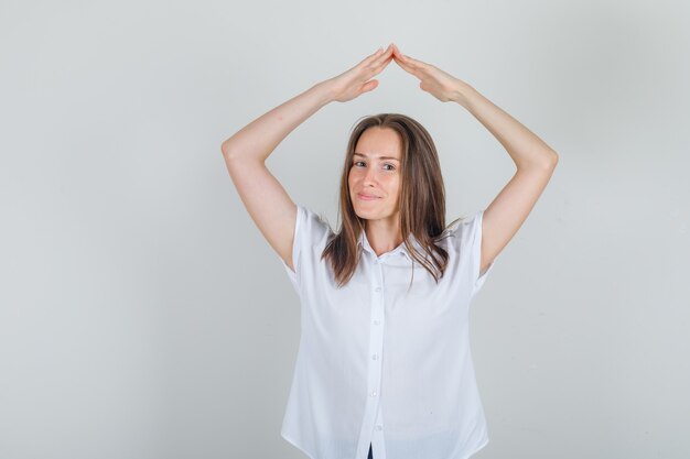 Mujer joven haciendo signo de techo de la casa por encima de la cabeza con camisa blanca y mirando alegre