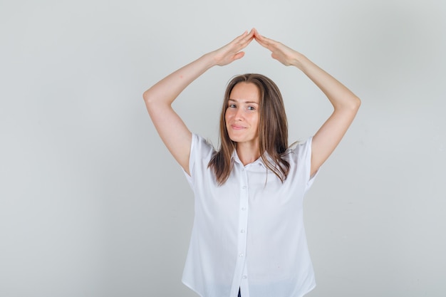 Mujer joven haciendo signo de techo de la casa por encima de la cabeza con camisa blanca y mirando alegre