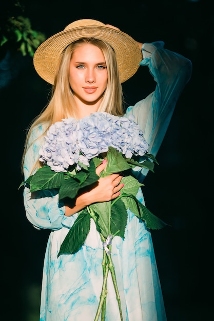 Foto gratuita mujer joven con hedragea buquet azul en vestido azul de pie en el jardín verde al atardecer