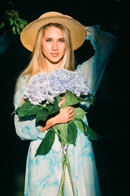 Mujer joven con hedragea buquet azul en vestido azul de pie en el jardín verde al atardecer