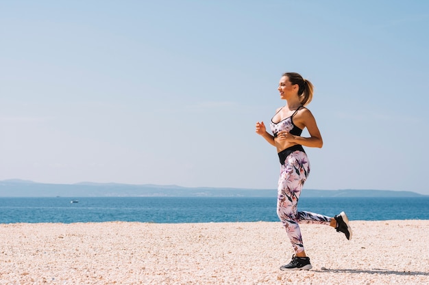 Mujer joven hermosa en el desgaste de la aptitud que corre en la playa cerca del mar