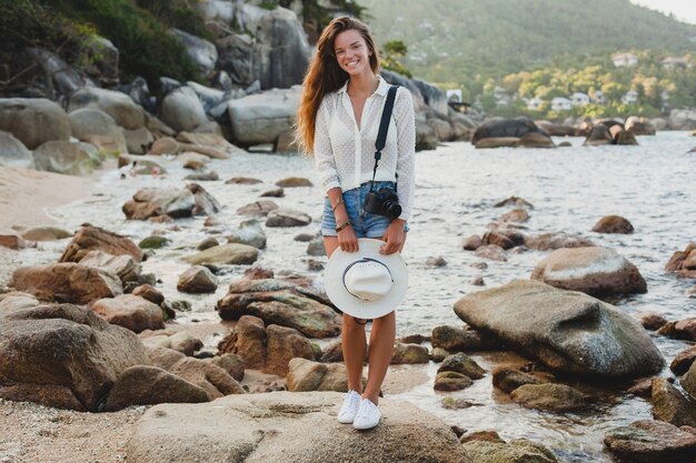 Mujer joven hermosa hipster en vacaciones de verano en asia, relajándose en la playa tropical, cámara de fotos digital, estilo boho casual, paisaje de mar, cuerpo delgado y bronceado, viajar solo