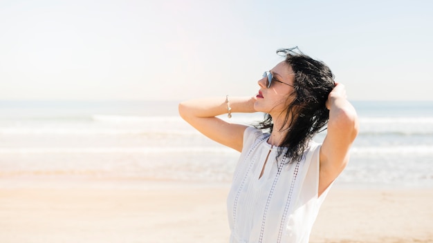 Foto gratuita mujer joven hermosa con la mano en su cabeza que se coloca cerca del mar en la playa