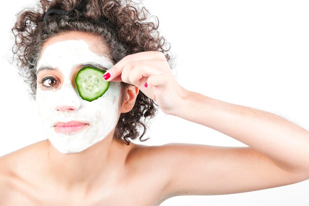 Mujer joven hermosa con la mascarilla que lleva a cabo la rebanada del pepino sobre su ojo