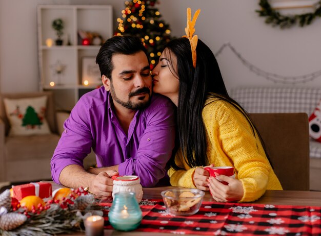 Mujer joven y hermosa pareja besando a hombre sentado en la mesa con tazas de té feliz en el amor en la habitación decorada de Navidad con árbol de Navidad en el fondo