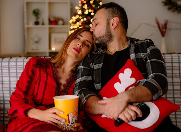Mujer joven y hermosa pareja durmiendo en el hombro de su novio feliz sentado en un sofá con un cubo de palomitas de maíz viendo la televisión juntos en la habitación decorada con árbol de Navidad en la pared