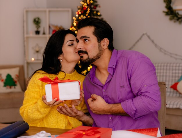 Mujer joven y hermosa pareja homding un regalo y besando a su novio en la habitación decorada de Navidad con árbol de Navidad en el fondo