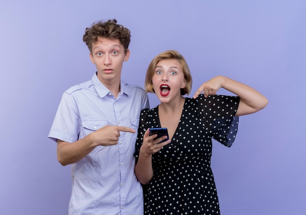 Mujer joven hermosa pareja sosteniendo el teléfono inteligente y apuntando con su dedo al teléfono inteligente con su novio parado sobre la pared azul