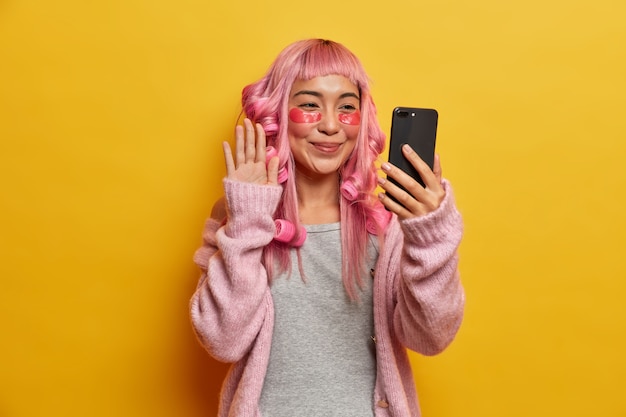 Mujer joven hermosa positiva se somete a procedimientos de belleza, usa rulos en el cabello teñido de rosa, aplica almohadillas de colágeno debajo de los ojos, hace selfie con teléfono inteligente