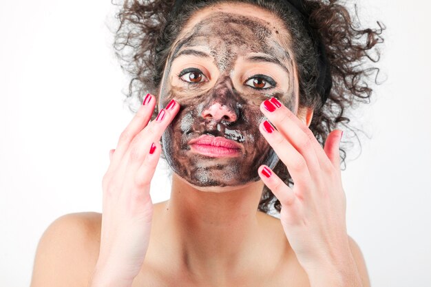 Mujer joven hermosa que aplica la mascarilla negra con sus dedos contra el fondo blanco