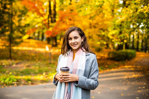 Mujer joven hermosa que bebe el café para llevar en parque en otoño.