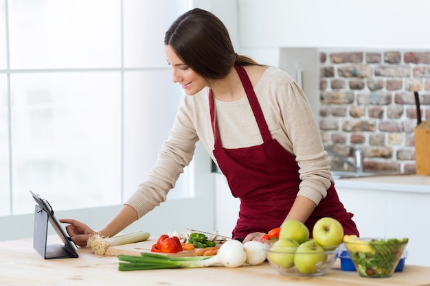 Mujer joven hermosa que cocina y que usa la tableta digital en la cocina.