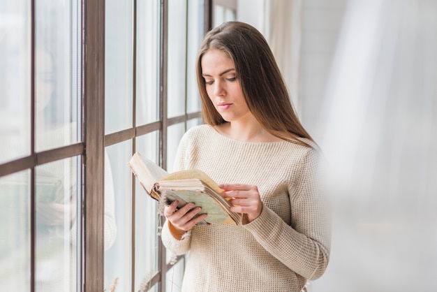 Mujer joven hermosa que se coloca cerca del libro de lectura de la ventana