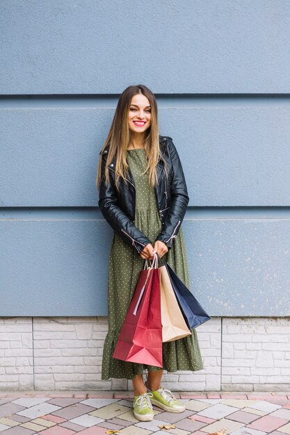 Mujer joven hermosa que se coloca delante de la pared que sostiene bolsos de compras coloridos