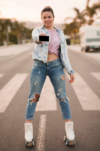 Mujer joven hermosa que se coloca en el patín de ruedas que muestra el teléfono móvil en el camino