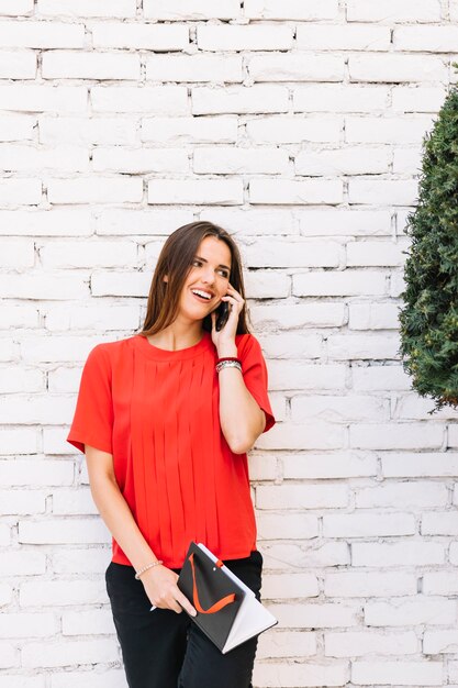 Mujer joven hermosa que habla en el teléfono celular delante de la pared de ladrillo