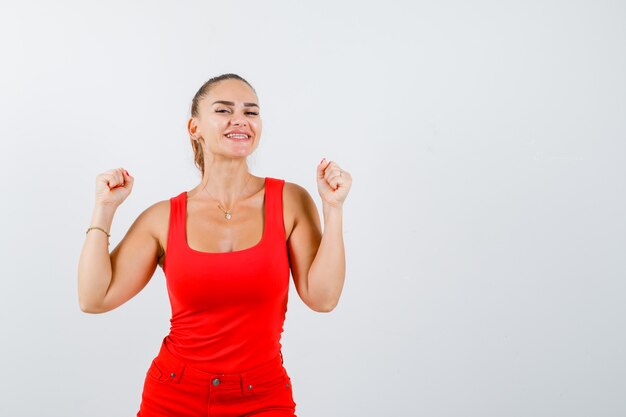 Mujer joven hermosa que muestra el gesto del ganador en la camiseta sin mangas roja y que parece feliz, vista frontal.