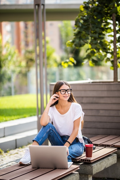 Mujer joven hermosa que se sienta en el banco que sostiene el café mientras que usa la computadora portátil al aire libre.