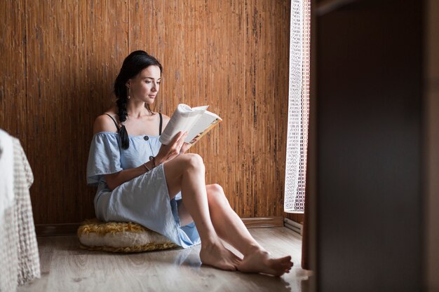 Mujer joven hermosa que se sienta cerca del libro de lectura de la ventana