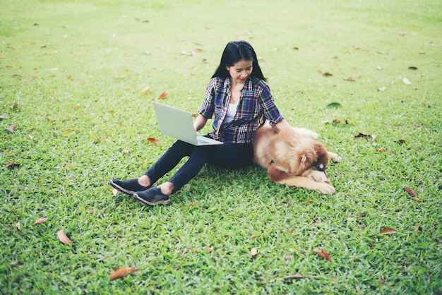 Mujer joven hermosa que usa la computadora portátil con su pequeño perro en un parque al aire libre. Estilo de vida.