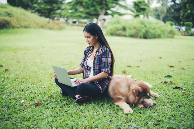 Mujer joven hermosa que usa la computadora portátil con su pequeño perro en un parque al aire libre. Retrato de estilo de vida