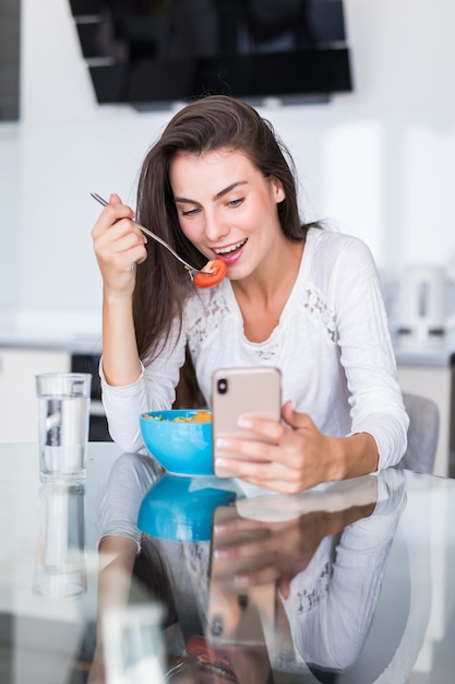 Mujer joven hermosa que usa el teléfono celular mientras que hace la ensalada en la cocina. Comida sana. ensalada de vegetales. Dieta. Estilo de vida saludable. Cocinar en casa.