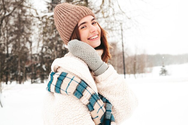 Mujer joven hermosa sonriente hipster en ropa de abrigo de moda y bufanda. Mujer despreocupada posando en la calle en el parque. Modelo puro positivo divirtiéndose en la nieve. Disfrutando de los momentos invernales