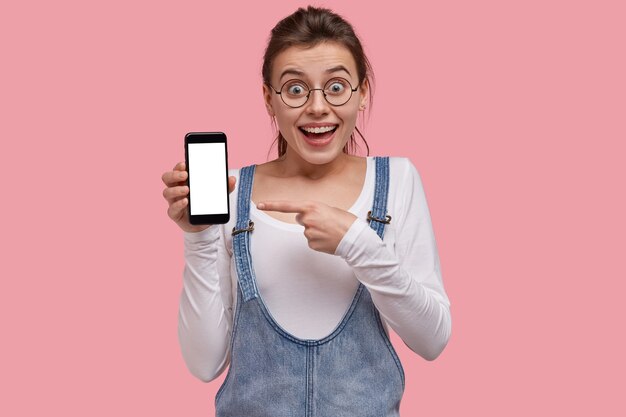 Mujer joven hermosa sorprendida indica en la pantalla en blanco del teléfono inteligente, muestra espacio para su información, vestida con traje de moda