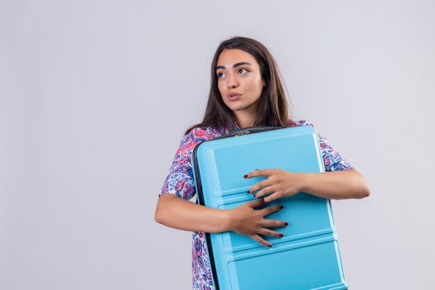 Mujer joven hermosa viajera abrazando la maleta azul mirando a un lado con expresión positiva en la cara de pie sobre fondo blanco.