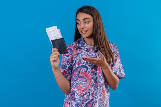 Mujer joven hermosa viajera con pasaporte con billetes apuntando con el brazo de la mano a ellos mirando positivo y feliz sonriendo de pie sobre fondo azul.