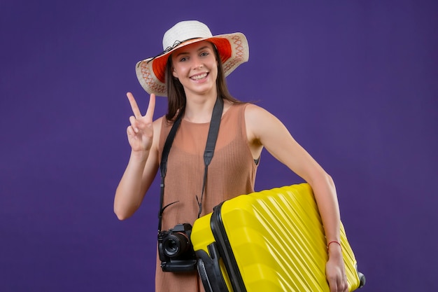 Mujer joven hermosa viajera con sombrero de verano con maleta amarilla y cámara positiva y feliz sonriendo alegremente mostrando el signo de la victoria o el número dos de pie sobre fondo púrpura