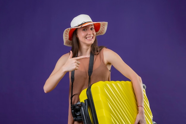 Foto gratuita mujer joven hermosa viajera con sombrero de verano con maleta amarilla y cámara positiva y feliz sonriendo apuntando con el dedo hacia el lado de pie sobre fondo púrpura