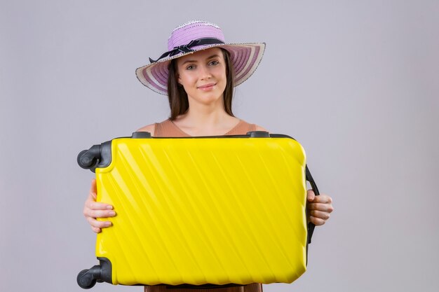 Mujer joven hermosa viajera con sombrero de verano con maleta amarilla sonriendo con cara feliz de pie sobre fondo blanco.