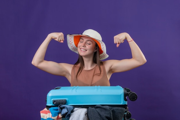 Mujer joven hermosa viajera con sombrero de verano con maleta llena de ropa levantando puños mostrando bíceps y fuerza optimista y feliz sonriendo de pie sobre fondo púrpura