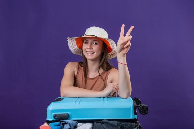 Mujer joven hermosa viajera con sombrero de verano con maleta llena de ropa positiva y feliz sonriendo alegremente optimista mostrando el signo de la victoria o el número dos de pie sobre fondo púrpura