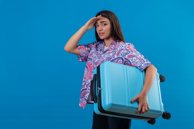Mujer joven hermosa viajera sosteniendo la maleta azul con aspecto cansado de pie con la mano en la cabeza sobre fondo azul.