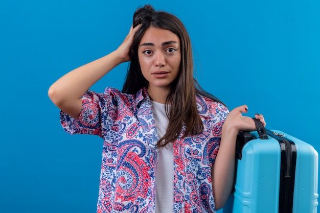 Mujer joven hermosa del viajero que sostiene la maleta azul que parece confundida con la mano en la cabeza para el error, recuerda el error, olvidó, concepto de la mala memoria sobre la pared azul aislada