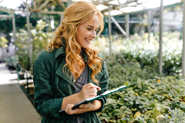 Mujer joven con hermoso cabello rubio y sonrisa suave, vestida con túnica verde con cinturón está trabajando en invernadero