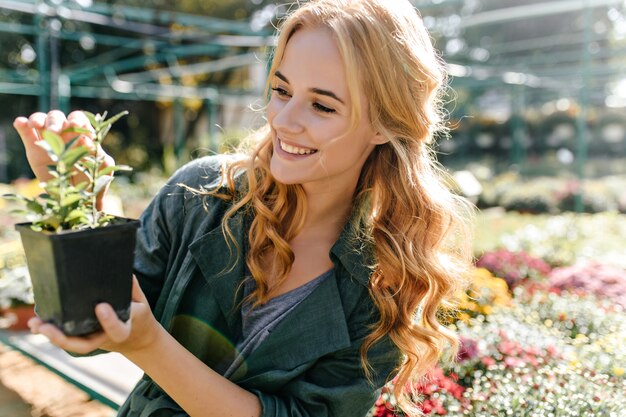 Mujer joven con hermoso cabello rubio y sonrisa suave, vestida con túnica verde con cinturón está trabajando en invernadero