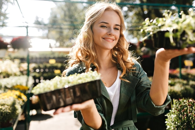 Mujer joven con hermoso cabello rubio y sonrisa suave, vestida con túnica verde con cinturón está trabajando en invernadero