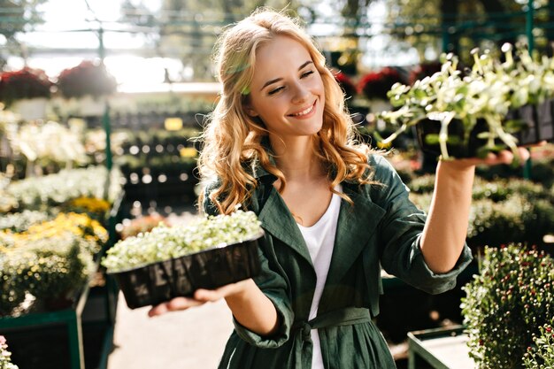 Mujer joven con hermoso cabello rubio y sonrisa suave, vestida con túnica verde con cinturón está trabajando en invernadero