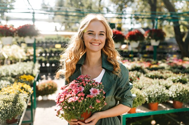 Mujer joven con hermoso cabello rubio y sonrisa suave, vestida con túnica verde con cinturón está trabajando en invernadero