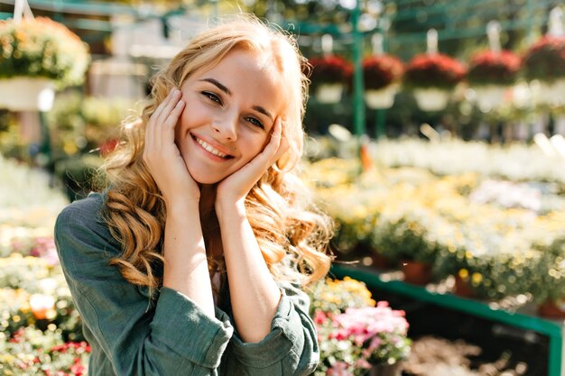 Mujer joven con hermoso cabello rubio y sonrisa suave, vestida con túnica verde con cinturón está trabajando en invernadero