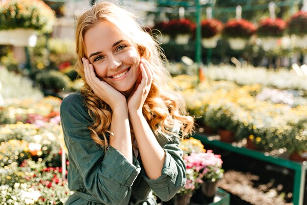 Mujer joven con hermoso cabello rubio y sonrisa suave, vestida con túnica verde con cinturón está trabajando en invernadero