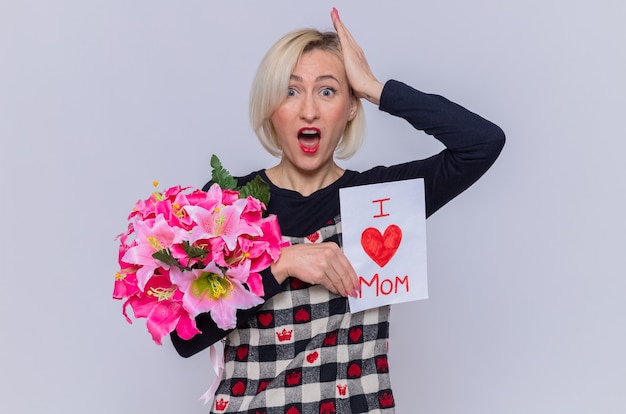 Mujer joven en un hermoso vestido sosteniendo una tarjeta de felicitación y un ramo de flores mirando al frente sorprendido con la mano en la cabeza celebrando el día de la madre de pie sobre una pared blanca