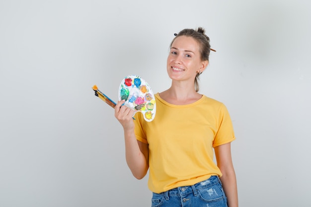 Mujer joven con herramientas de pintura en camiseta amarilla, pantalones cortos de jeans y aspecto alegre.