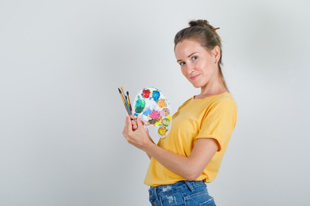Mujer joven con herramientas de pintura en camiseta amarilla, pantalones cortos de jeans y aspecto alegre.