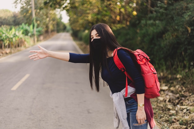 Mujer joven hippie con mascarilla haciendo autostop.