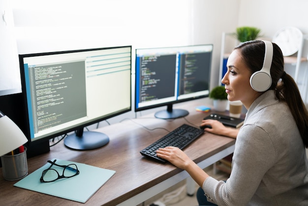 Mujer joven hispana con auriculares codificando una aplicación de software en la computadora en casa. Programador freelance escribiendo código